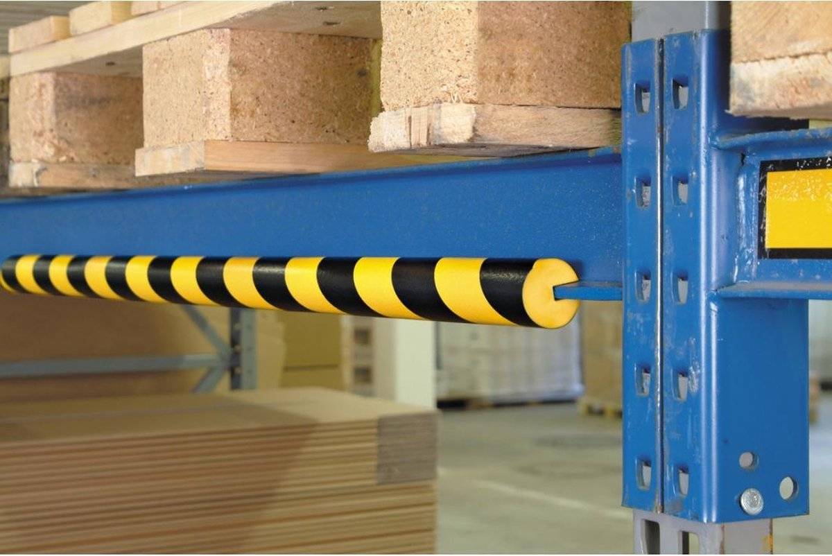 Ein blauer Metallträger mit einer gelb und schwarz gestreiften Schaumstoffschutzvorrichtung ist unter Holzregalen montiert. Holzpaletten sind im Hintergrund sichtbar.