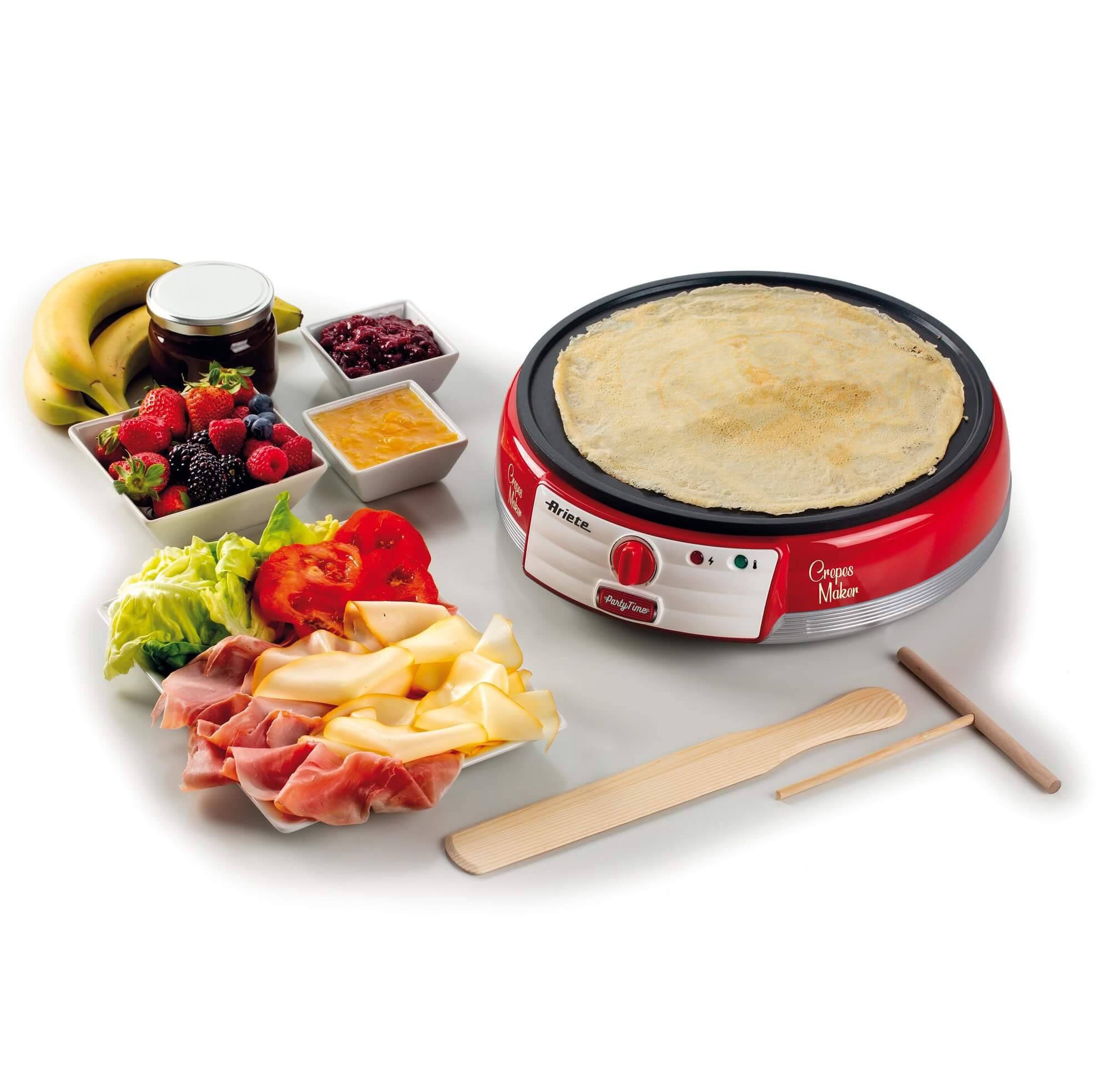 Elektrische Crêpes-Maker mit einer zubereiteten Crêpe, serviert mit Obst, Marmeladen, Salat, Käse und Schinken auf weißem Hintergrund.