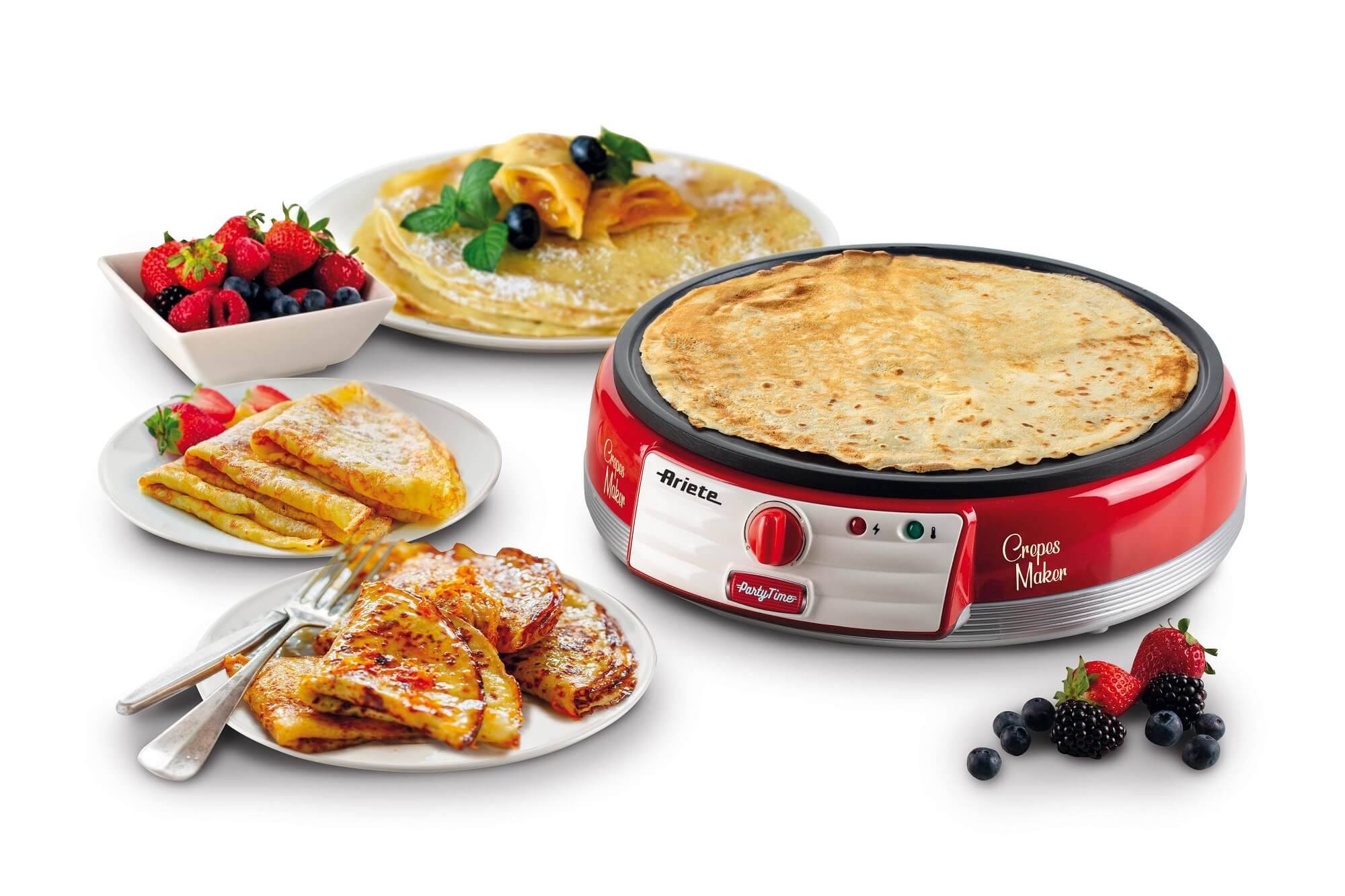 Roter elektrischer Crêpes-Maker mit einer gekochten Crêpe, umgeben von Tellern mit gefalteten Crêpes, die mit Obst, Butter und Sauce auf weißem Hintergrund belegt sind.