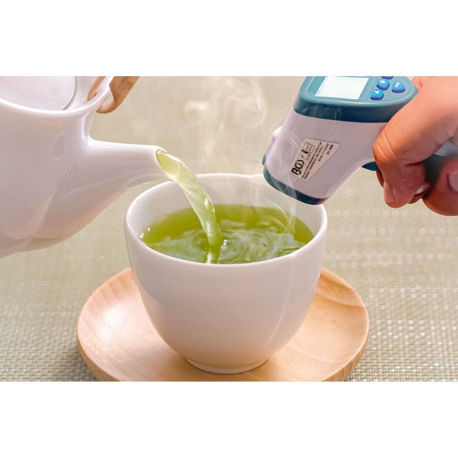 Heißes Wasser wird in eine weiße Tasse gegossen, während eine Person mit einem Thermometer die Temperatur des Getränks misst.