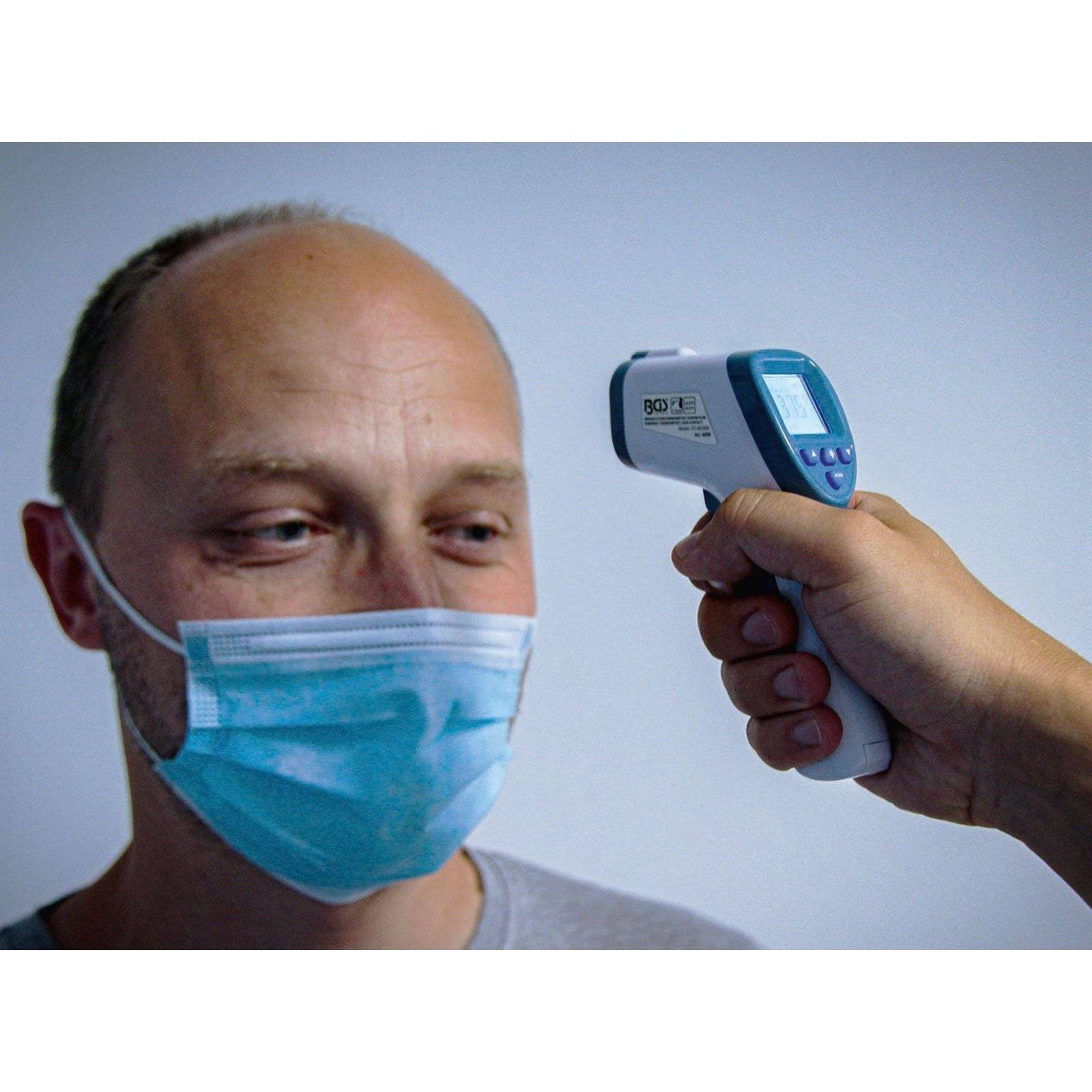 Eine Person mit Gesichtsmaske lässt sich von einer anderen Person mit einem digitalen Stirnthermometer die Temperatur messen.