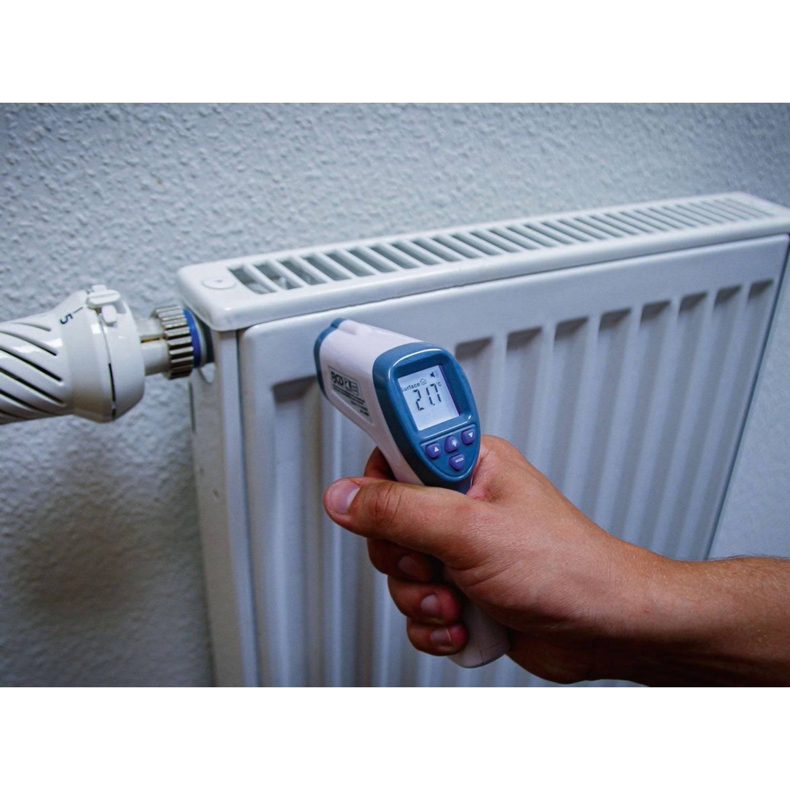 Eine Hand hält einen Infrarot-Thermometer, der ‚27,7°C' in der Nähe eines Heizkörpers anzeigt und damit die Oberflächentemperatur der Heizeinheit misst.