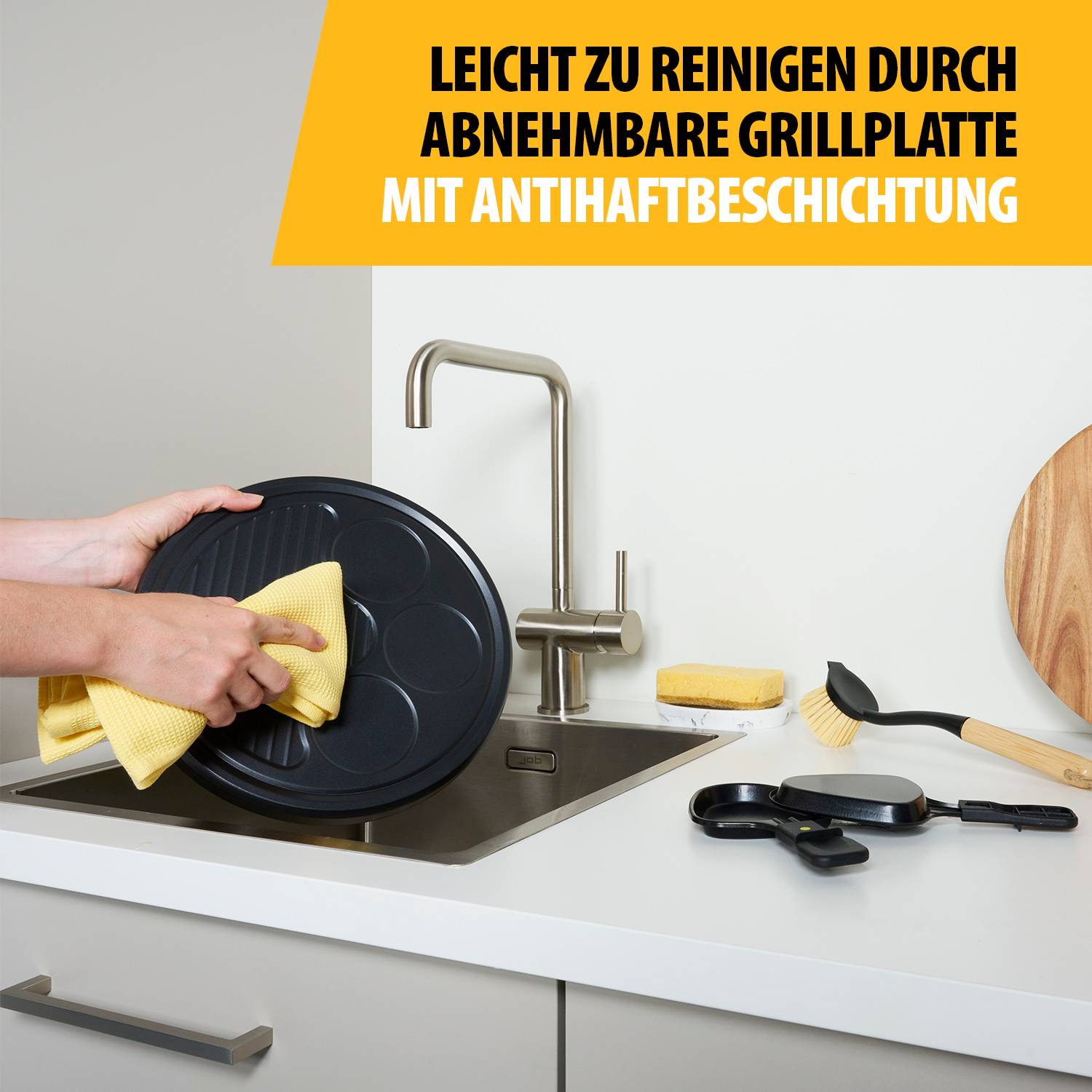 Hände reinigen eine abnehmbare Grillplatte mit einem Tuch an einer Küchenspüle; Text sagt „LEICHT ZU REINIGEN DURCH ABNEHMBARE GRILLPLATTE MIT ANTIHAFTBESCHICHTUNG"