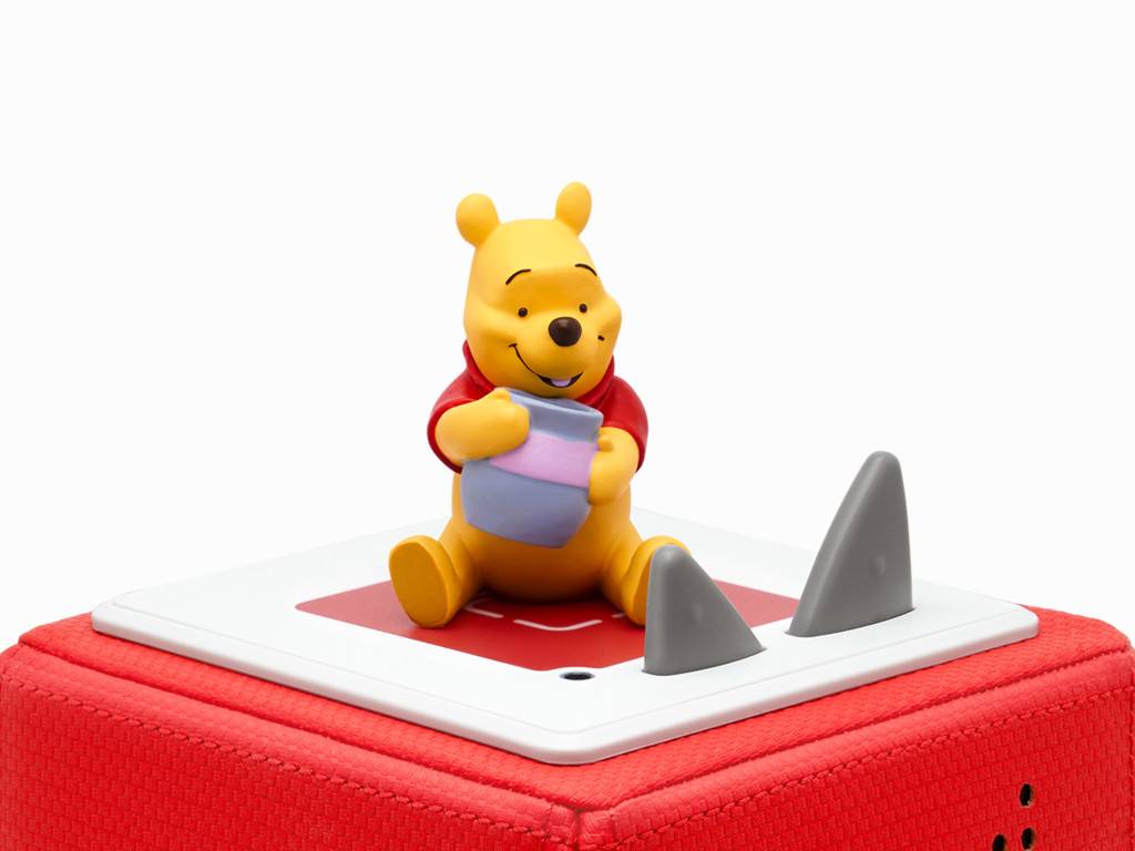 Eine Winnie-Puuh-Figur sitzt auf einem weißen, tablettenähnlichen Gerät auf einem roten Kissen, mit zwei grauen kegelförmigen Objekten daneben.