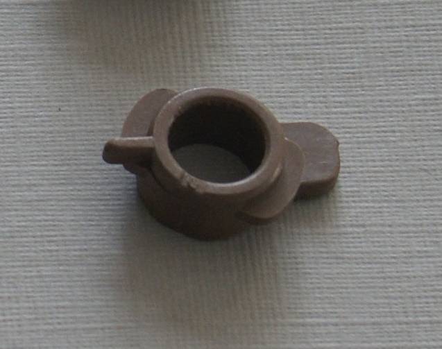 Ein kleines, braunes Kunststoffbauteil mit einer kreisförmigen Öffnung und hervorstehenden Laschen, auf einer strukturierten, hellfarbigen Oberfläche platziert.