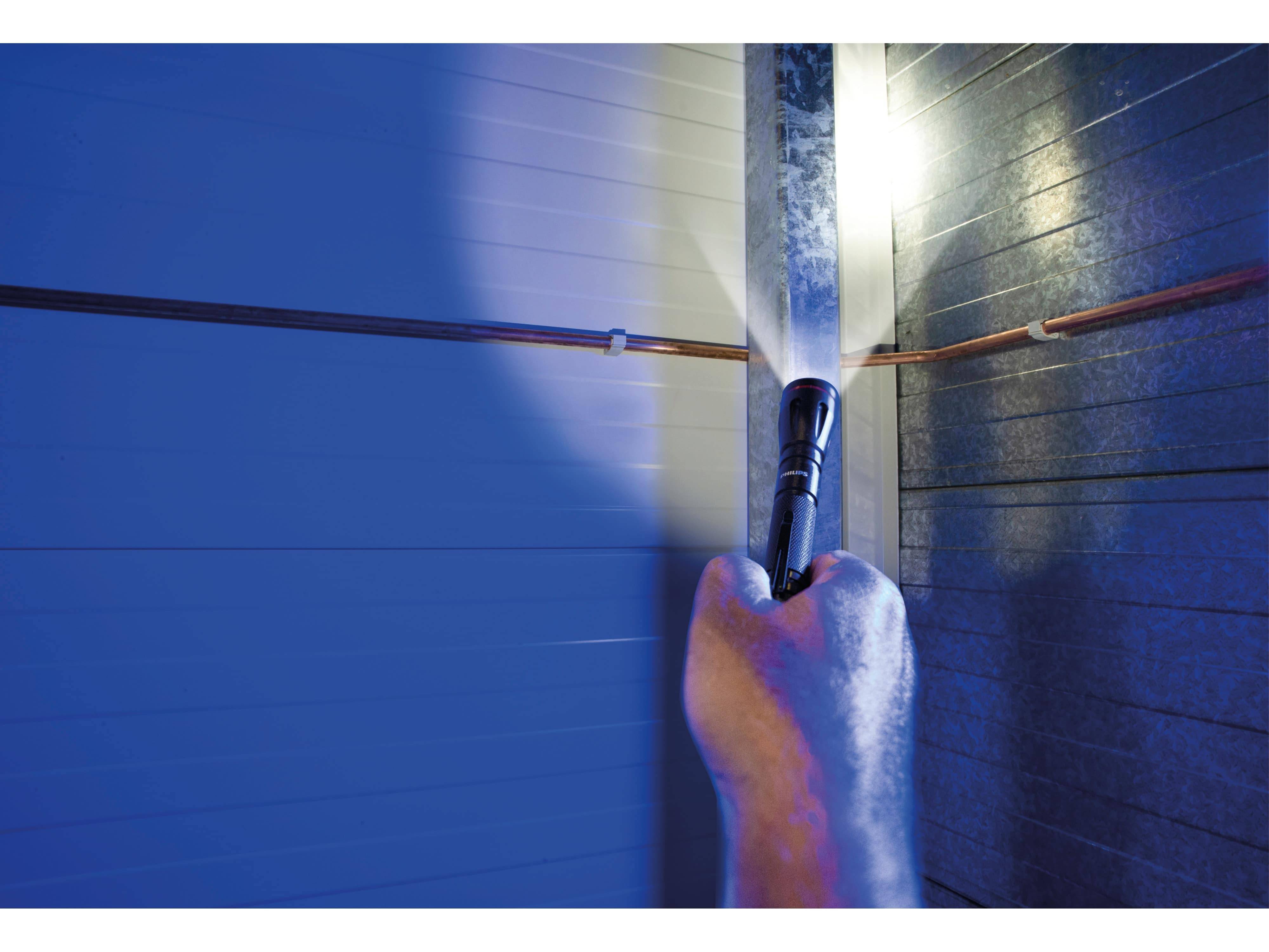 Eine Hand, die eine Taschenlampe in einem schwach beleuchteten Technikraum hält und ein metallisches Rohr sowie die umgebende Wand beleuchtet.