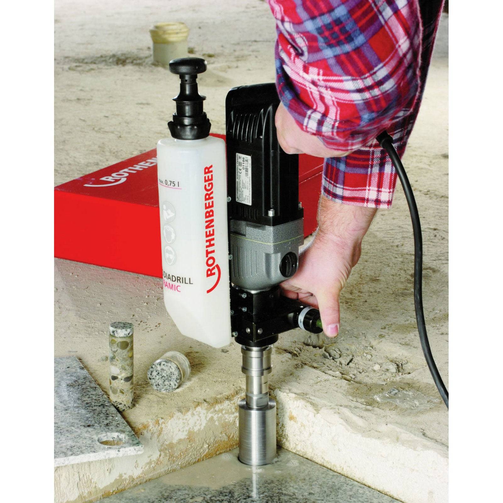 Person mit einer elektrischen Kernbohrmaschine, die ein Loch in eine Betonoberfläche bohrt. Die Bohrmaschine weist sichtbare Markierung und einen zylindrischen Bohrer auf.