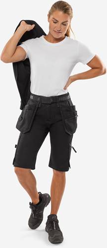 Eine Person mit einem weißen Hemd und schwarzen Arbeitsshorts mit mehreren Taschen, die mit einer Hand auf der Hüfte steht und eine lässige Kleidung trägt.