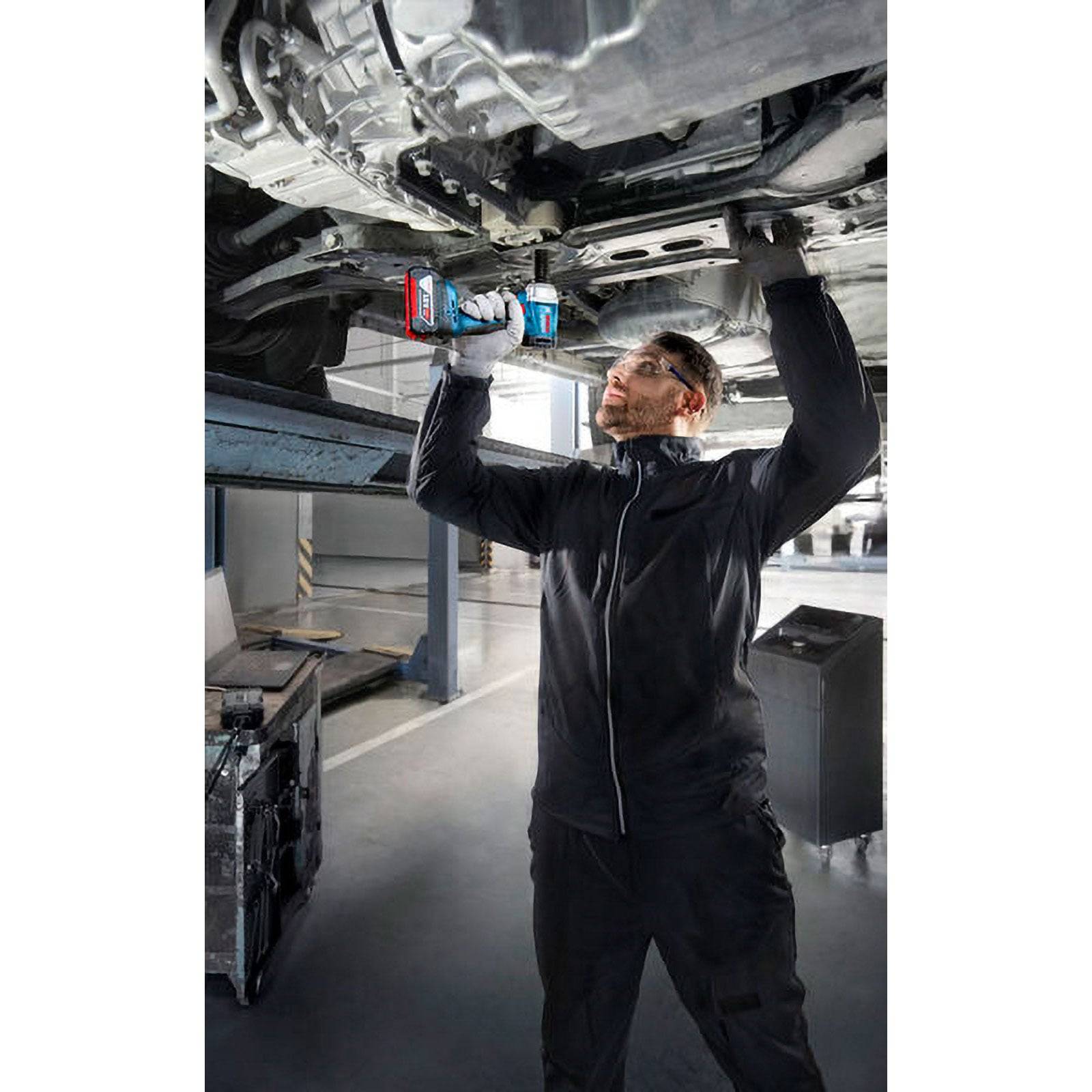 Ein Mechaniker in schwarzer Arbeitskleidung benutzt ein Elektrowerkzeug, um die Unterseite eines Fahrzeugs in einer Autoservicewerkstatt zu bearbeiten.