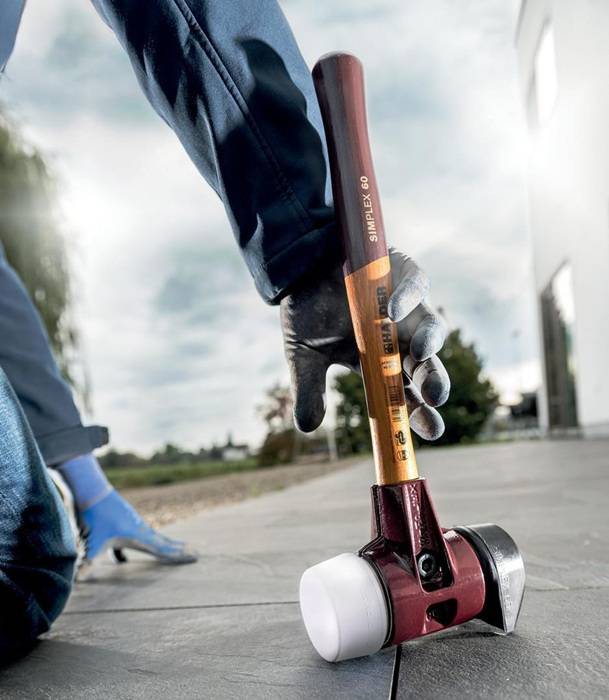 Eine Person mit Handschuhen verwendet einen Hammer mit einem weißen Gummimallet-Kopf auf einer Steinoberfläche im Freien und konzentriert sich auf präzise Platzierung.