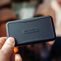 Hand hält ein kleines schwarzes GLYPH-Gerät, ein rechteckiges Gadget mit abgerundeten Ecken vor einem unscharfen Innenraum-Hintergrund.