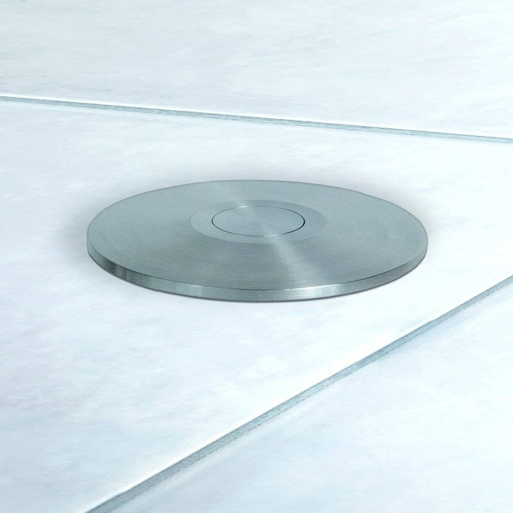 Eine runde, metallische Scheibe, die auf einem gefliesten Boden befestigt ist, möglicherweise als Ablaufabdeckung dienend. Sie ist zentral positioniert, was auf Funktionalität hindeutet.