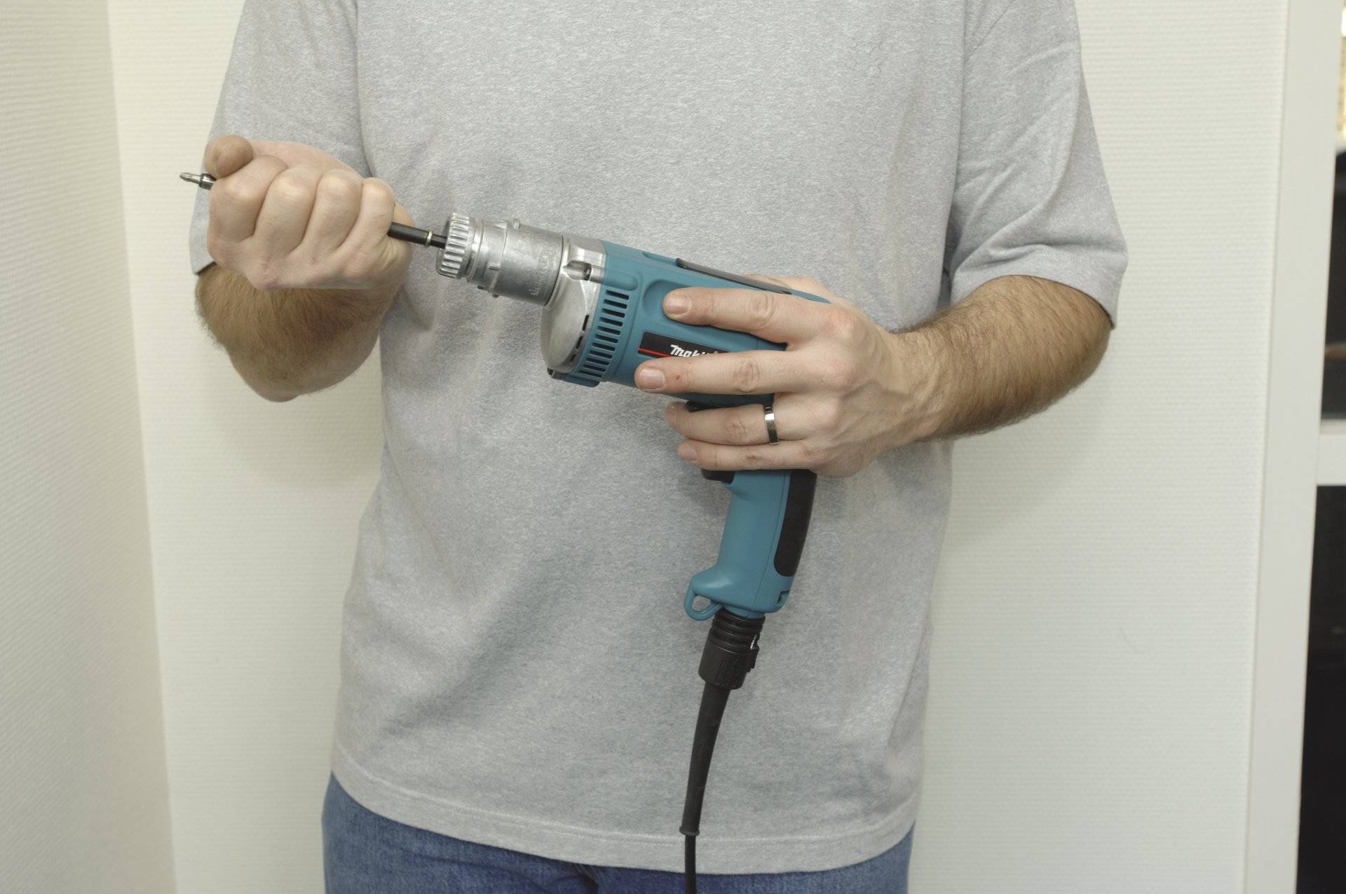 Eine Person, die einen elektrischen Bohrer hält und ein graues T-Shirt und Jeans trägt. Der Bohrer ist blau mit einem schwarzen Kabel. Die Person stellt den Bohraufsatz ein.