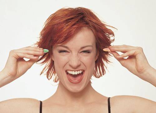 Eine Person mit kurzen roten Haaren, die lächelt und ihre Hände nahe der Ohren hebt. Ihr Gesichtsausdruck ist fröhlich und energiegeladen.