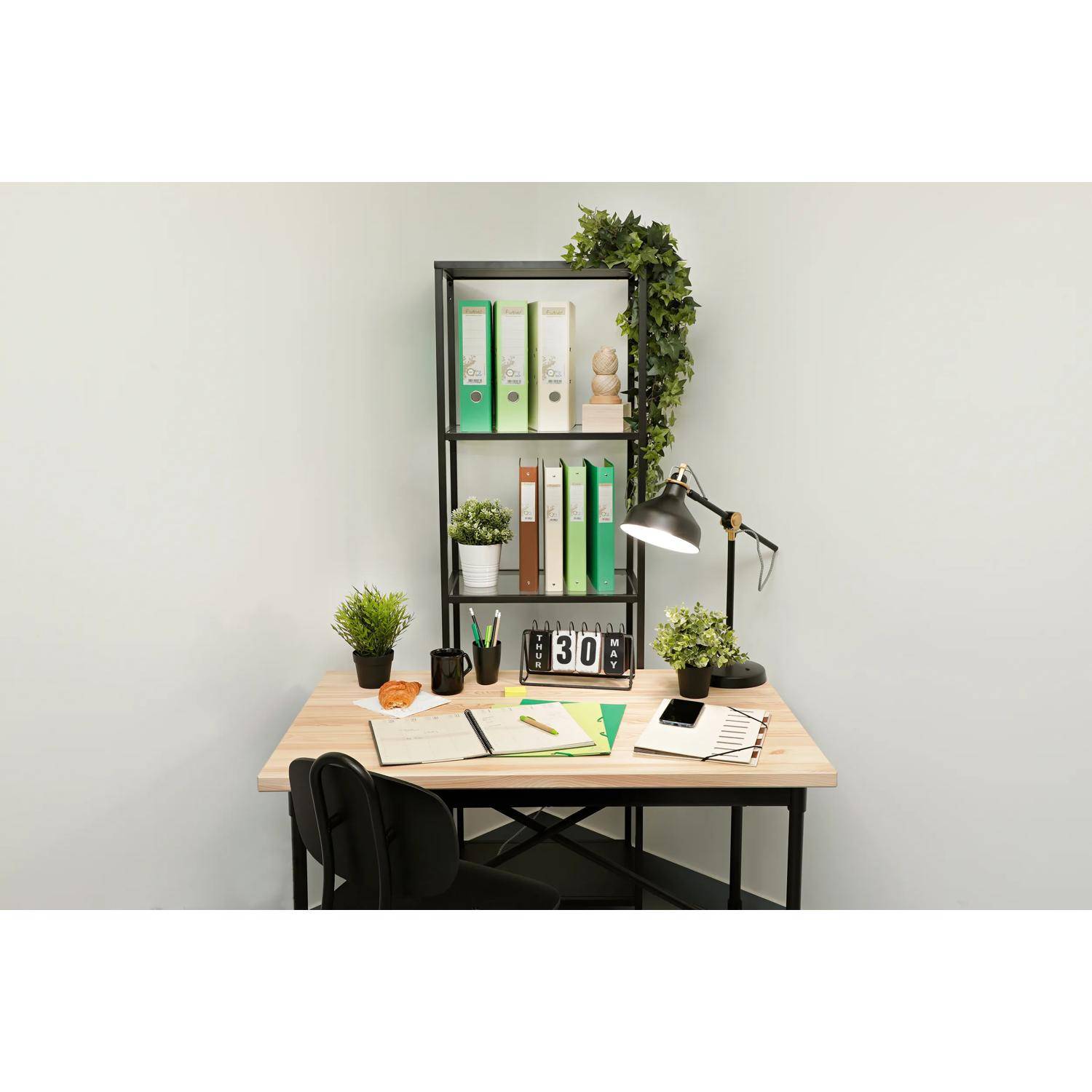 Minimalistischer Arbeitsplatz im Homeoffice mit schwarzem Stuhl, Regalen mit Ordnern und Pflanzen, Schreibtischlampe, Uhr und Notizbüchern vor einer schlichten Wand.