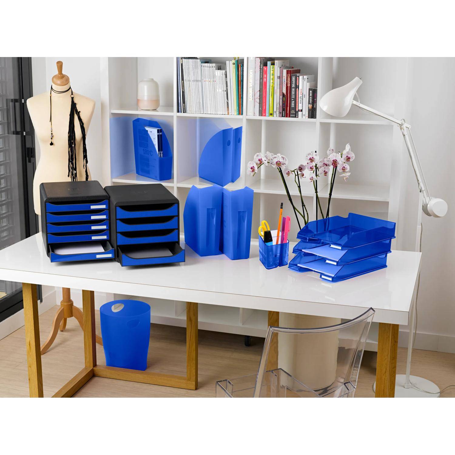 Organisierter Homeoffice-Schreibtisch mit blauen Aktenablagen, Mappen, Stifthalter, Bücherregalen, Schneiderpuppe und Schreibtischlampe auf einem weißen Tisch.