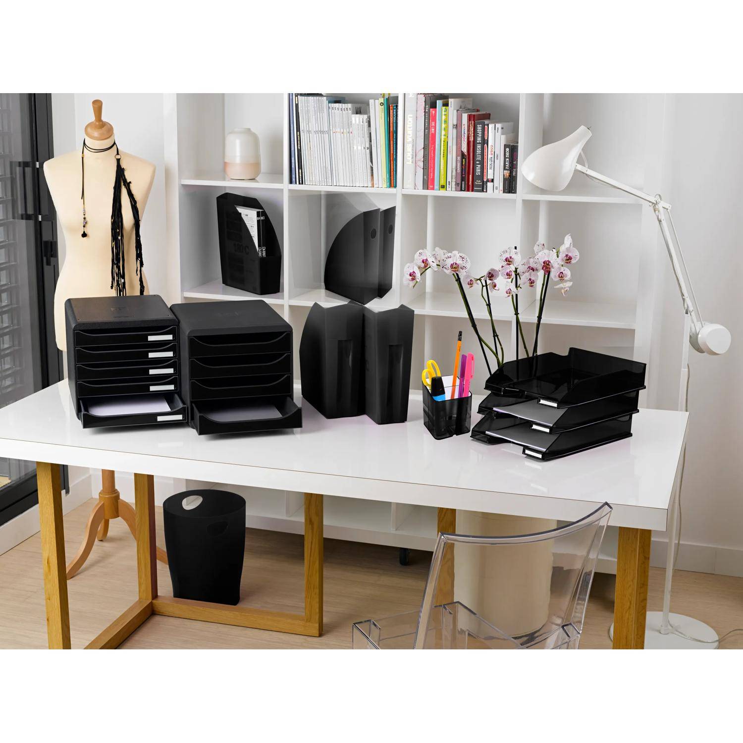 Homeoffice-Schreibtisch mit schwarzen Ablagefächern, Ordnern, Büchern, Stiften und einer Orchidee auf einem weißen Tisch neben einer Schneiderpuppe und einer Lampe.