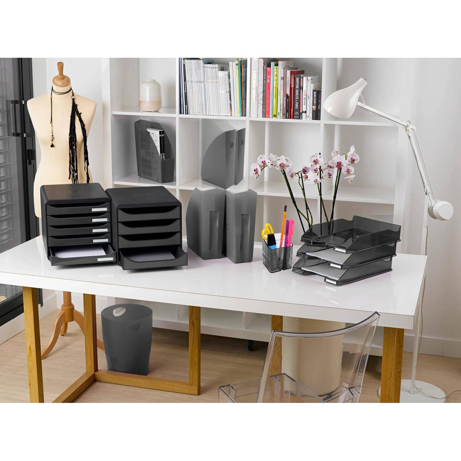 Ein ordentlicher Schreibtisch im Homeoffice mit schwarzen Ablageschalen, Büromaterialien, Orchideenblüten, Bücherregalen und einer Schneiderpuppe im Hintergrund.