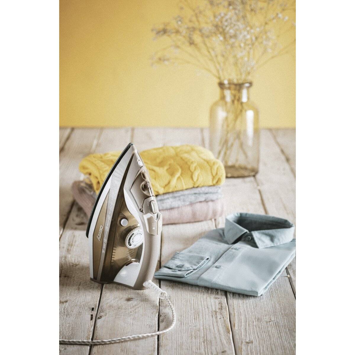 Ein Bügeleisen steht auf einem Holztisch neben gefalteter Kleidung, darunter ein blaues Hemd und ein gelbes Handtuch. Eine Vase mit getrockneten Blumen steht im Hintergrund.