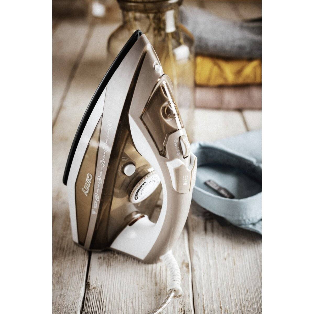 Ein Dampfbügeleisen wird aufrecht auf einer Holzoberfläche neben gefalteter Kleidung und einer Glasflasche platziert, was auf eine Wäsche- oder Bügelumgebung hindeutet.