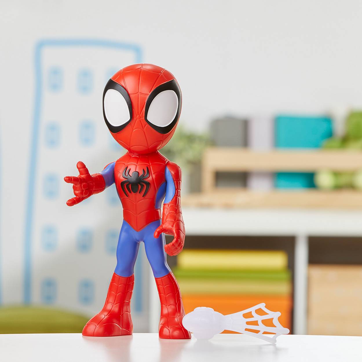 Spider-Man-Spielzeugfigur, die auf einer weißen Oberfläche steht, mit einem kleinen Spinnennetz-Teil daneben, in einer unscharfen Raumeinstellung.
