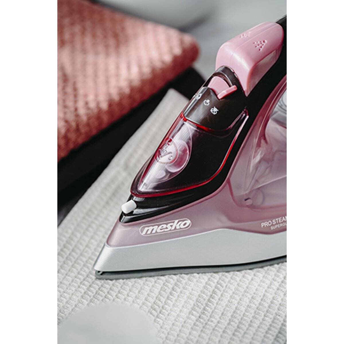 Ein Nahaufnahmebild eines rosa-schwarzen Dampfbügeleisens auf einem weißen strukturierten Stoff, mit einem roten gefalteten Handtuch im Hintergrund.