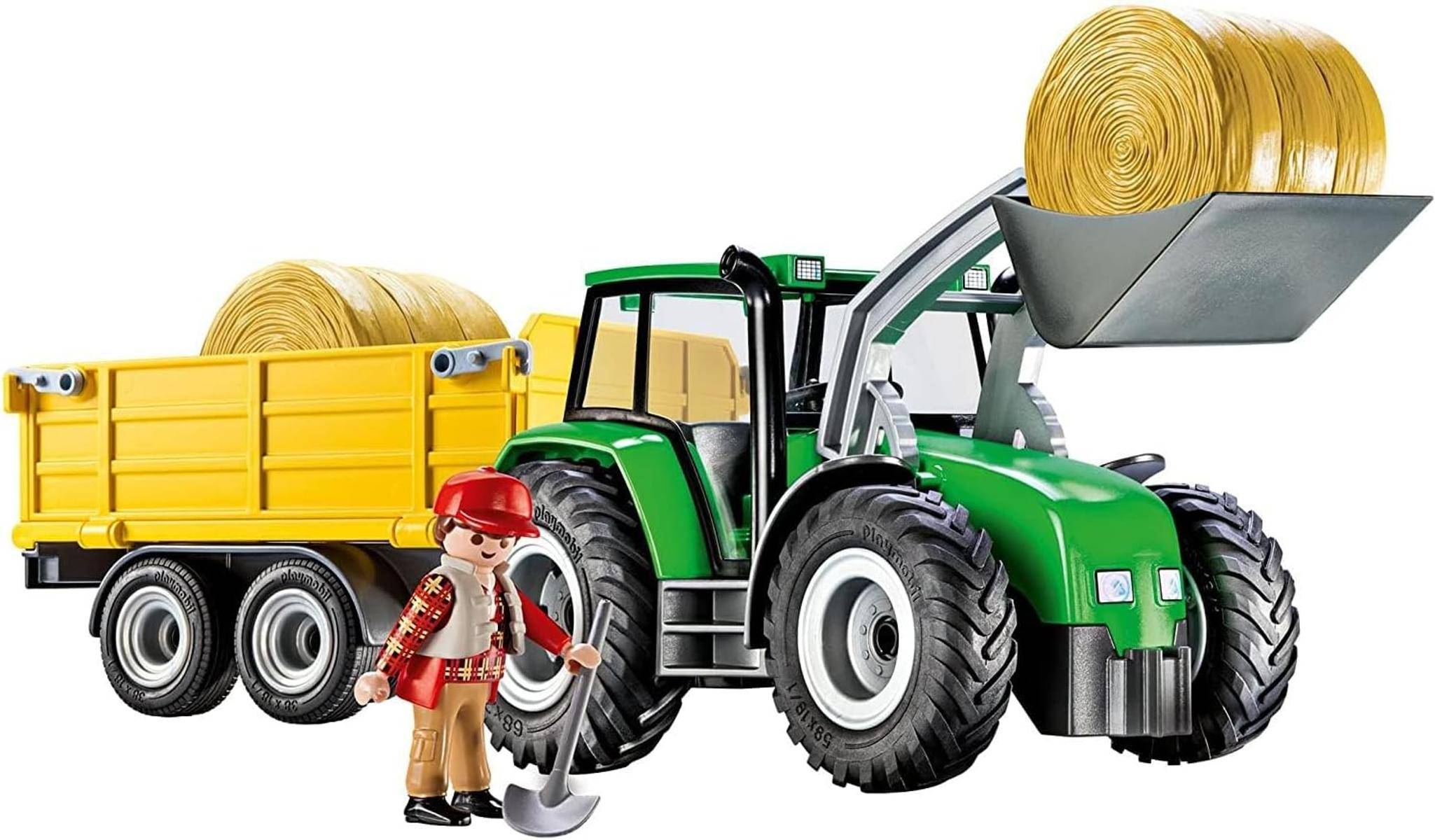 Grüner Spielzeugtraktor mit Frontlader, der Heuballen in einen gelben Anhänger lädt, mit einer Bauernfigur, die eine Heugabel hält.
