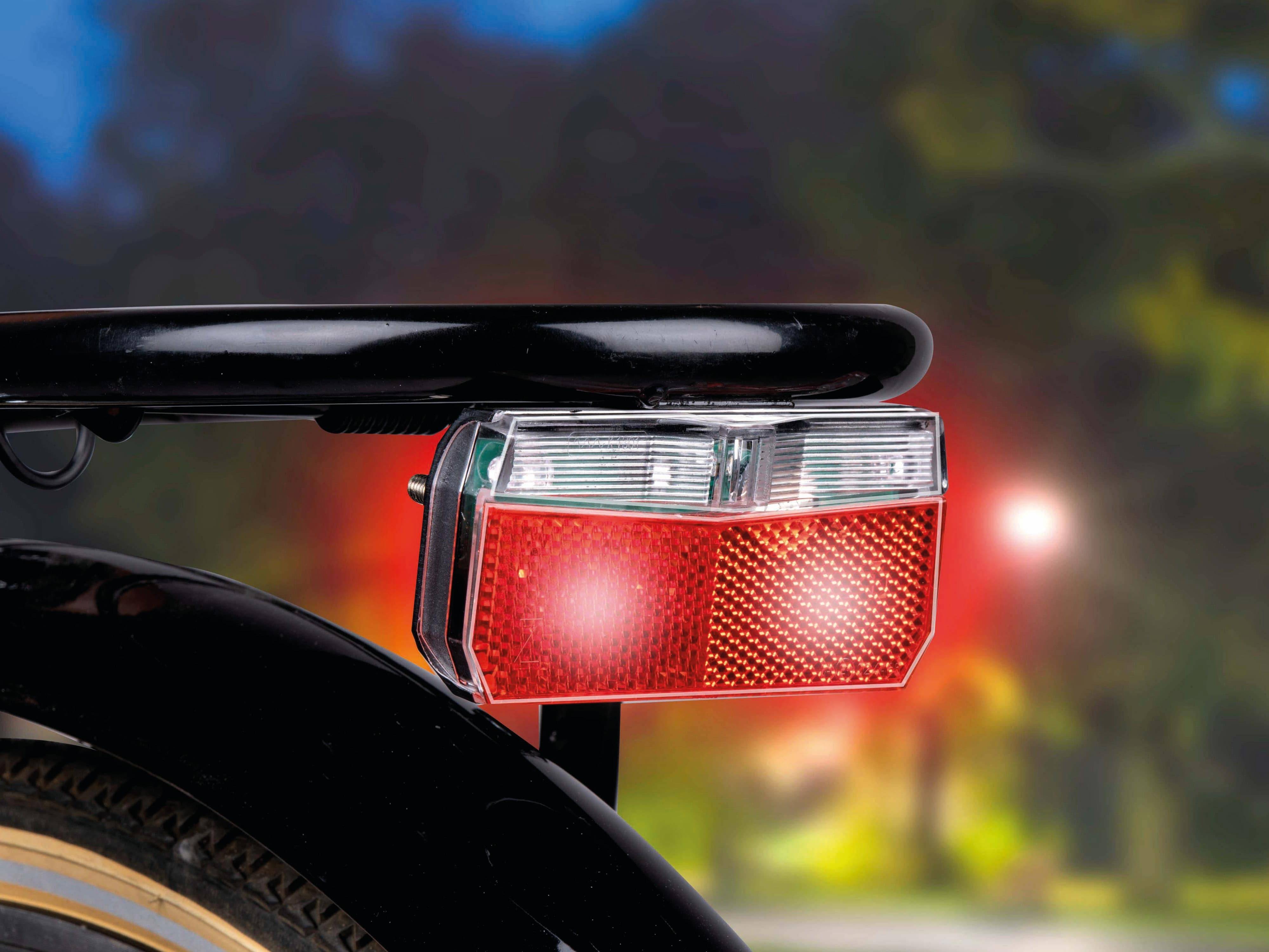 Rückansicht eines Fahrrads mit einem roten Reflektor und eingeschaltetem Rücklicht, was auf Nachtfahrten oder schwache Lichtverhältnisse hindeutet.