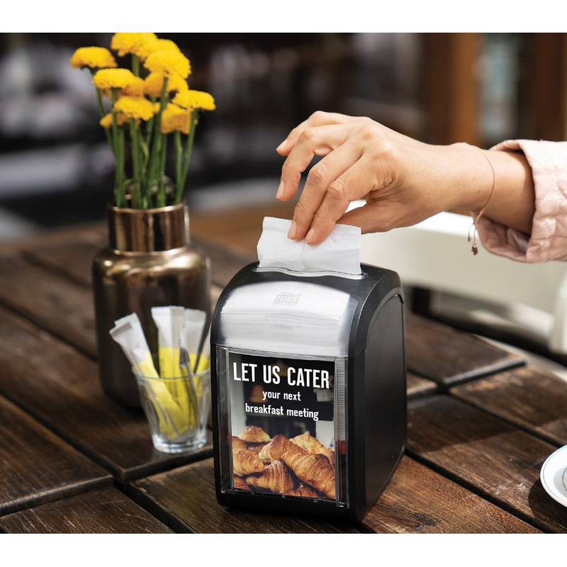 Eine Hand zieht eine Serviette aus einem Spender auf einem Holztisch, mit einem Schild, das die Aufschrift trägt: „Wir versorgen Ihre nächste Frühstücksbesprechung