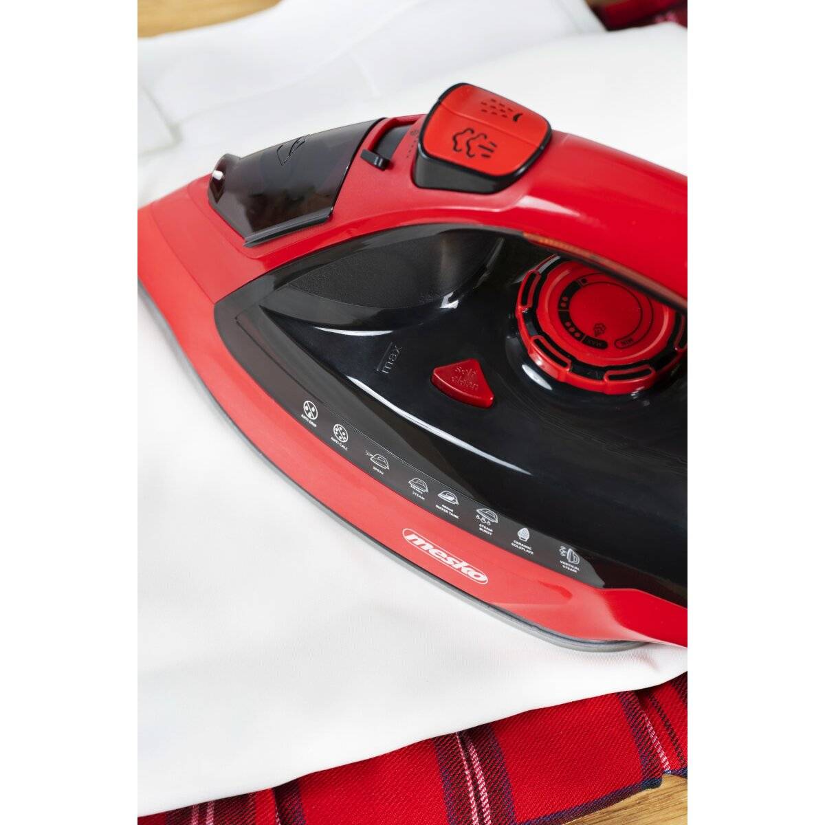 Ein rotes Elektrobügeleisen auf einem weißen Stoff platziert, über einem rot-schwarz gestreiften Tuch, mit sichtbarem Dampfreglerknopf und verschiedenen Einstellungen.