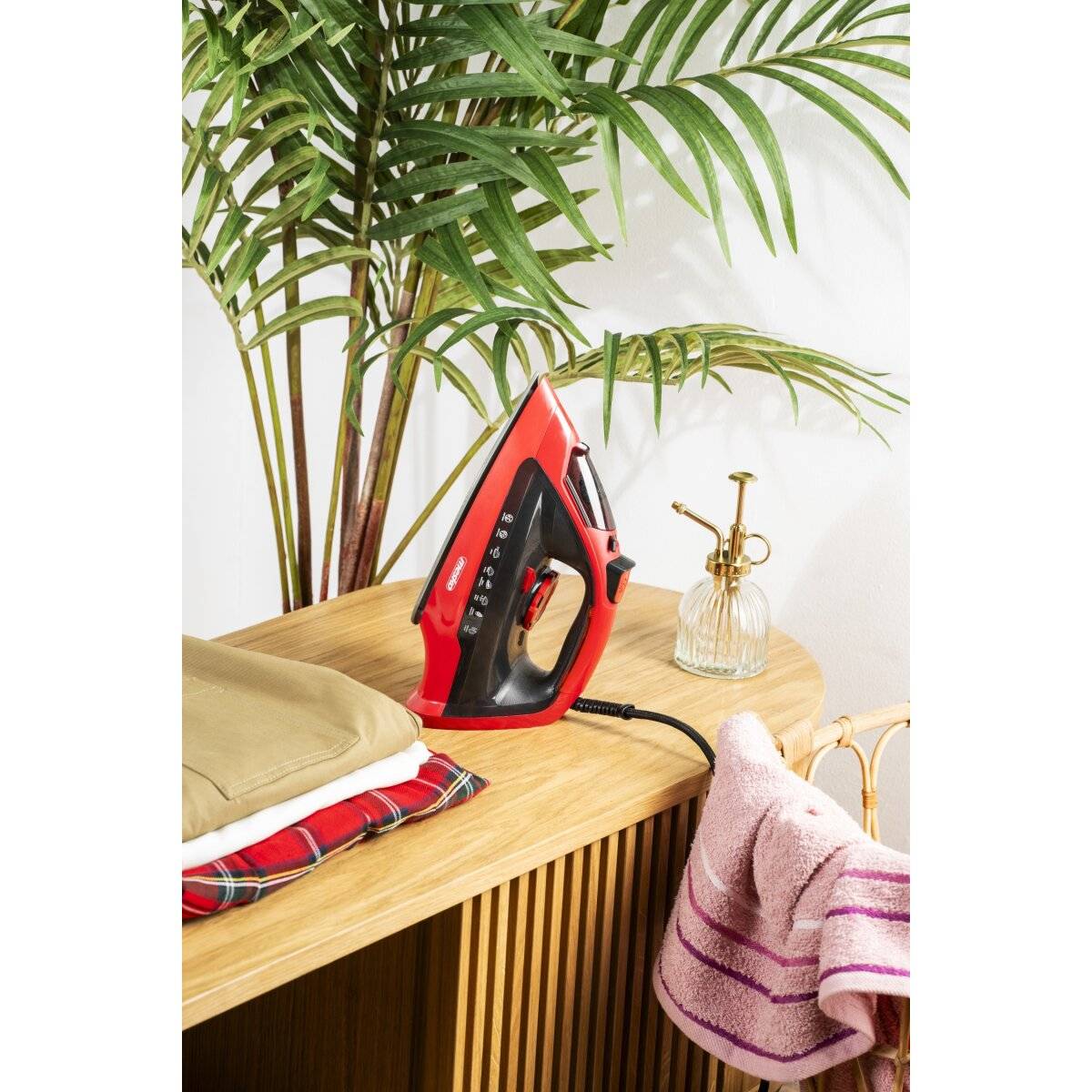 Ein rotes Dampfbügeleisen steht auf einem Holztisch neben gefalteter Kleidung und einer Palme. Eine Sprühflasche und Handtücher sind ebenfalls sichtbar.