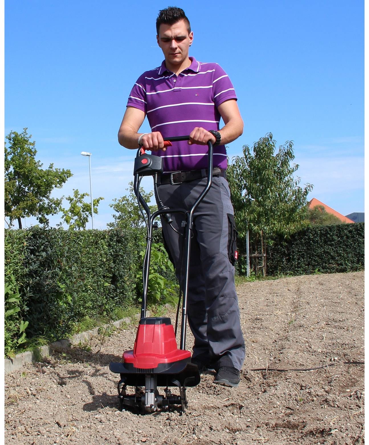 Eine Person bedient einen roten Gartenfräser in einem Garten und steht auf einem Stück Erde unter einem klaren blauen Himmel, mit grünen Hecken im Hintergrund.