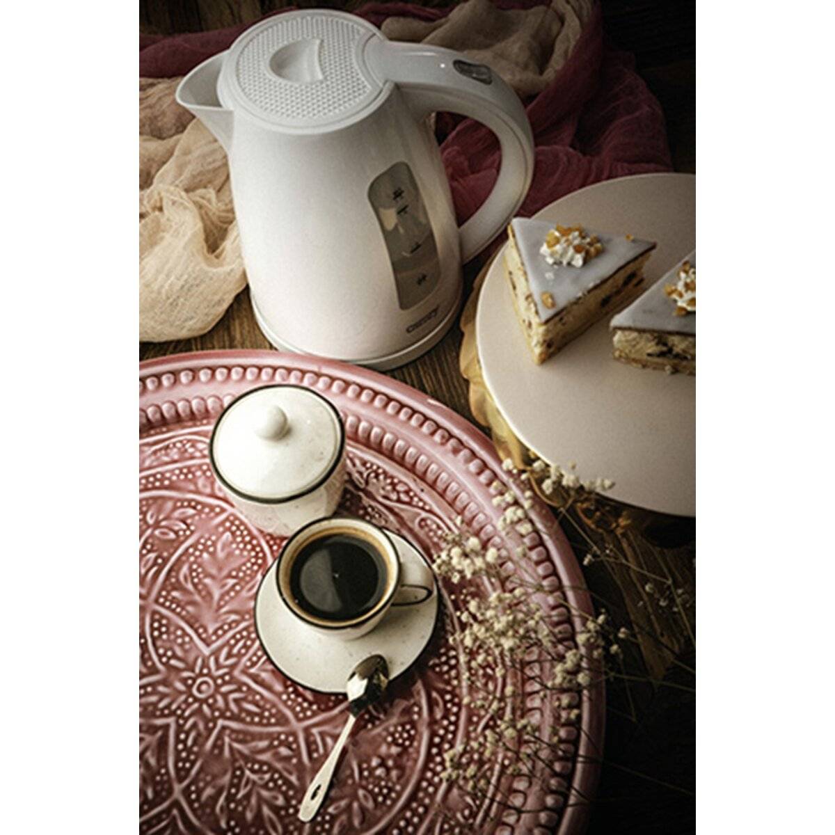 Ein weißer elektrischer Wasserkessel auf einem strukturierten rosa Tablett neben einer Tasse schwarzem Kaffee und einem Stück Kuchen auf einem weißen Teller, das eine gemütliche Atmosphäre schafft.