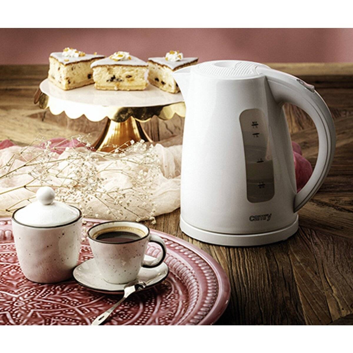 Ein weißer elektrischer Wasserkocher auf einem Holztisch neben zwei Tassen Kaffee auf einem dekorativen Tablett, mit einem Kuchen auf einem Ständer im Hintergrund.