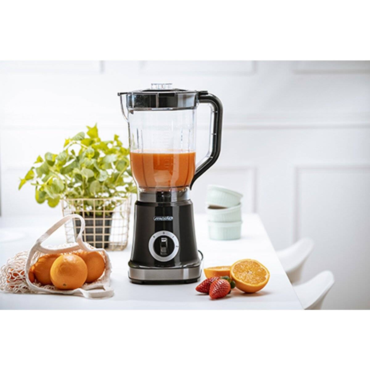 Ein mit orangefarbenem Smoothie gefüllter Mixer steht auf einer Küchentheke. In der Nähe befinden sich frische Orangen, Erdbeeren und eine grüne Pflanze.