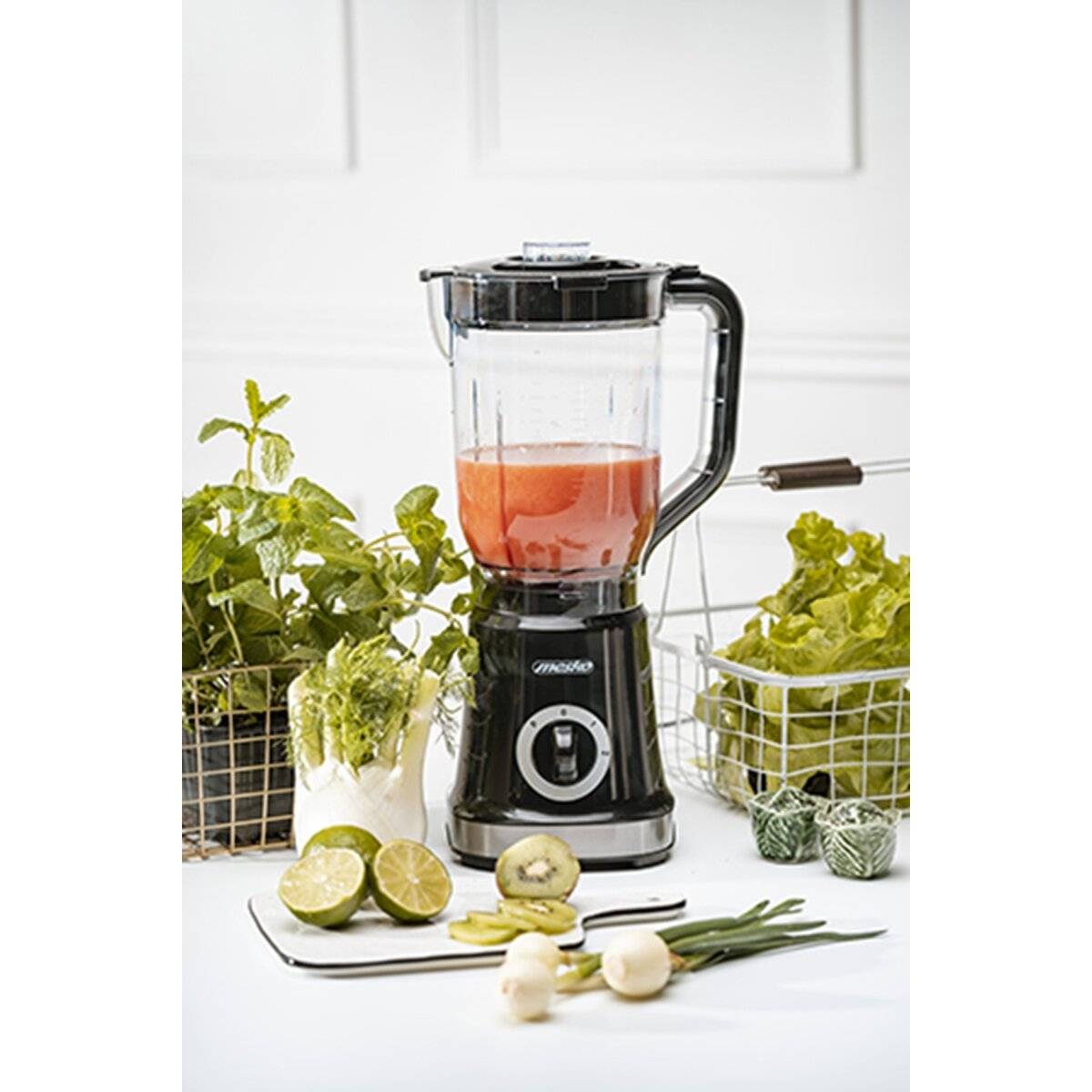 Ein schwarzer Mixer mit roter Flüssigkeit auf einer Küchentheke, umgeben von frischem Grünzeug, einer Limette und einer Zwiebel, was auf die Zubereitung einer gesunden Mahlzeit hindeutet.