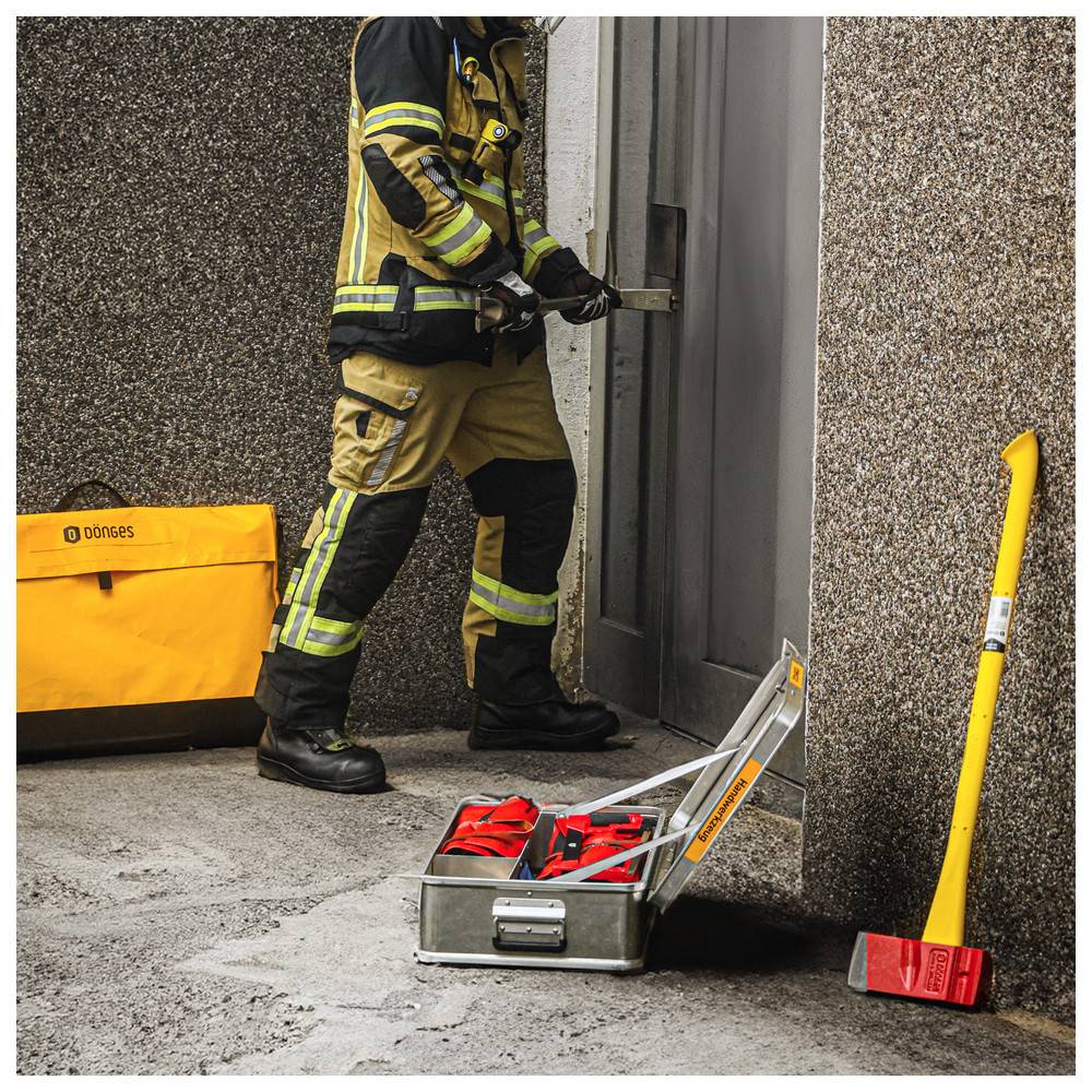 Ein Feuerwehrmann in Schutzausrüstung nutzt ein Werkzeug, um eine Tür zu öffnen. Eine gelbe Tasche, ein Metallwerkzeugkasten und eine gelbe Axt sind in der Nähe sichtbar und deuten auf eine Notfallreaktion hin.