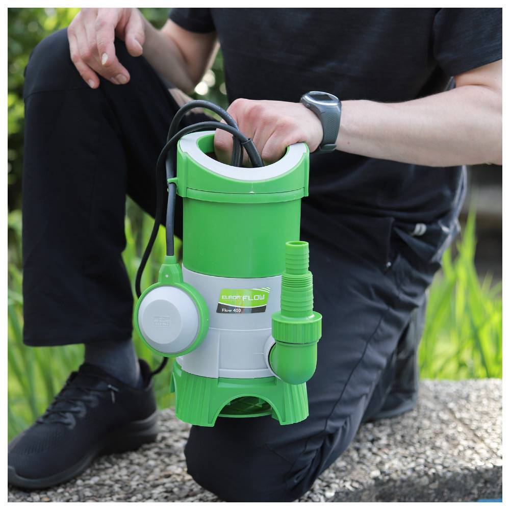 Person kniet auf Kies und hält eine grün-weiße Wasserpumpe mit schwarzem Kabel, trägt schwarze Hose und Schuhe, im Hintergrund eine Grasfläche.