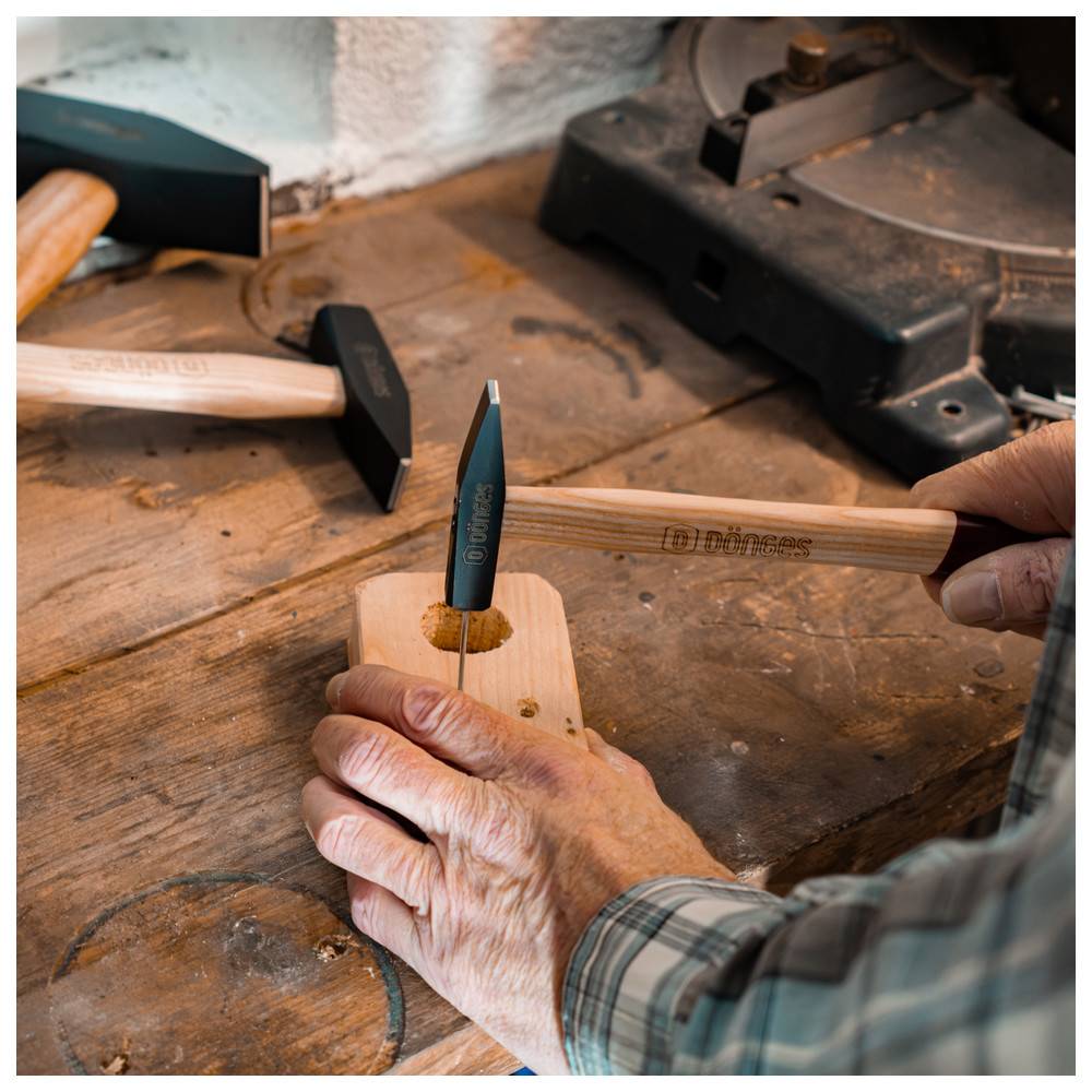 Eine Person verwendet einen Hammer und ein Locheisen an einem Holzblock mit Löchern. Ein weiterer Hammer liegt auf der Werkbank. Szene aus der Holzbearbeitung.