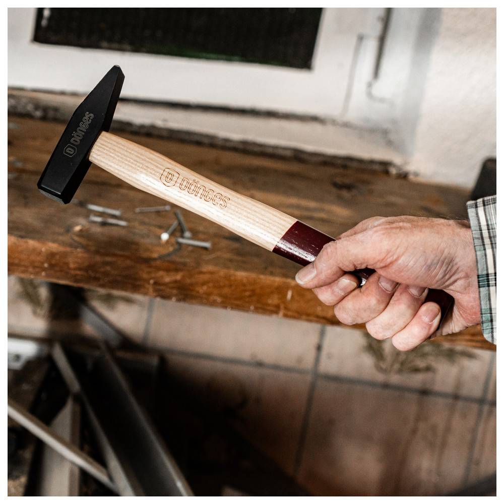 Eine Hand hält einen VÚMPS-Hammer mit einem schwarzen Kopf und einem Holzstiel. Mehrere Nägel liegen auf einer nahe gelegenen Holzoberfläche.