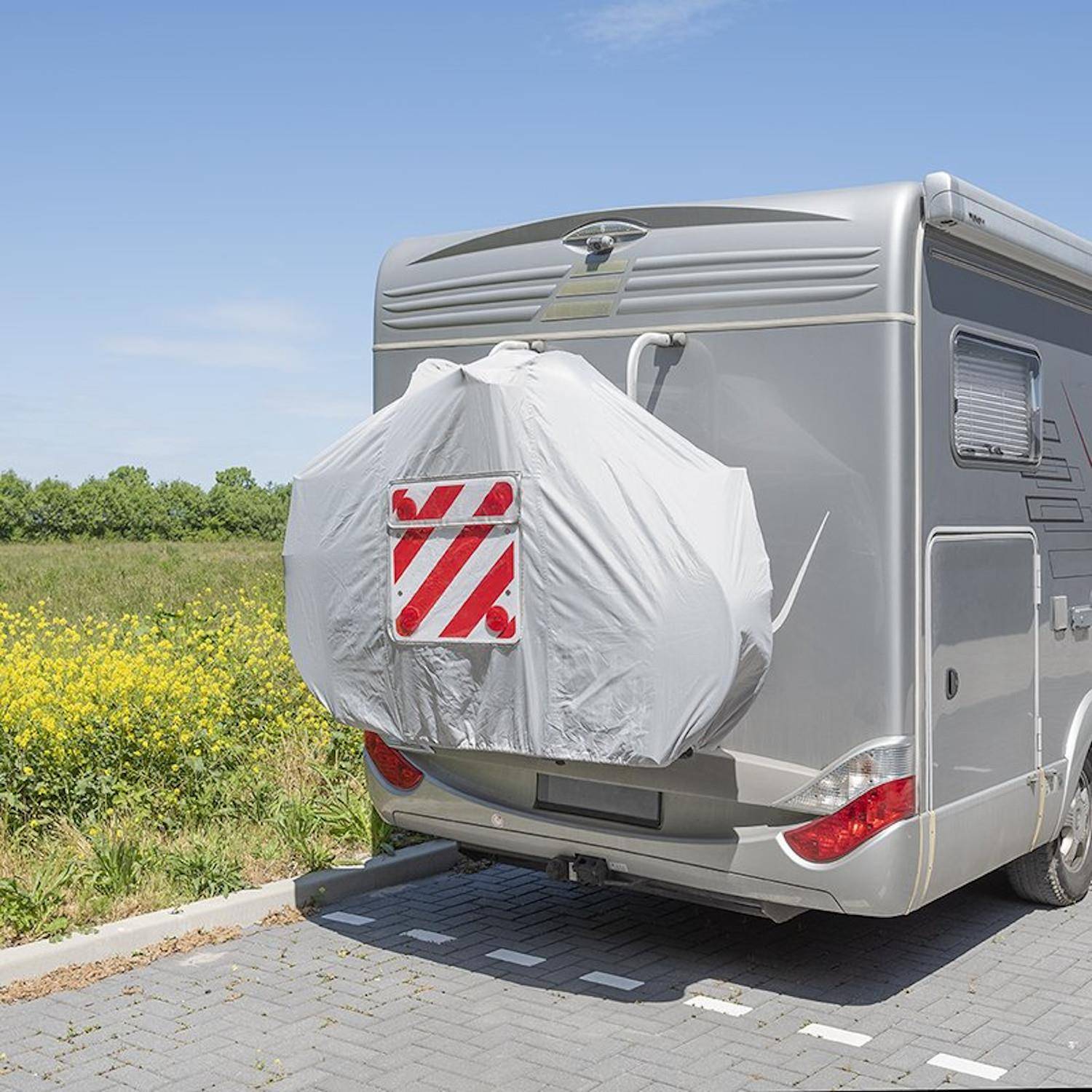 Ein silberner Wohnwagen, der auf einem gepflasterten Platz neben einem Feld mit gelben Blumen geparkt ist. Ein mit weißem Stoff bedeckter Reifen und ein rot-weißes Warnschild sind an der Rückseite angebracht.