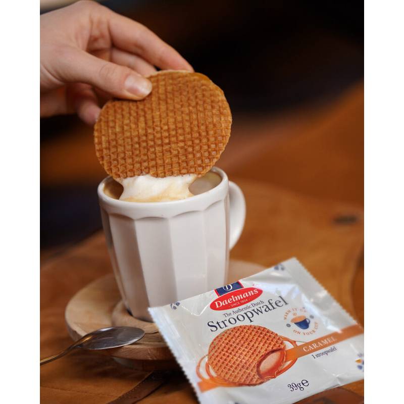 Eine Hand taucht eine Karamel-Stroopwafel über eine Tasse heißem Kaffee ein, mit einer verpackten Daelmans Stroopwafels-Packung und einem Löffel auf dem Tisch.