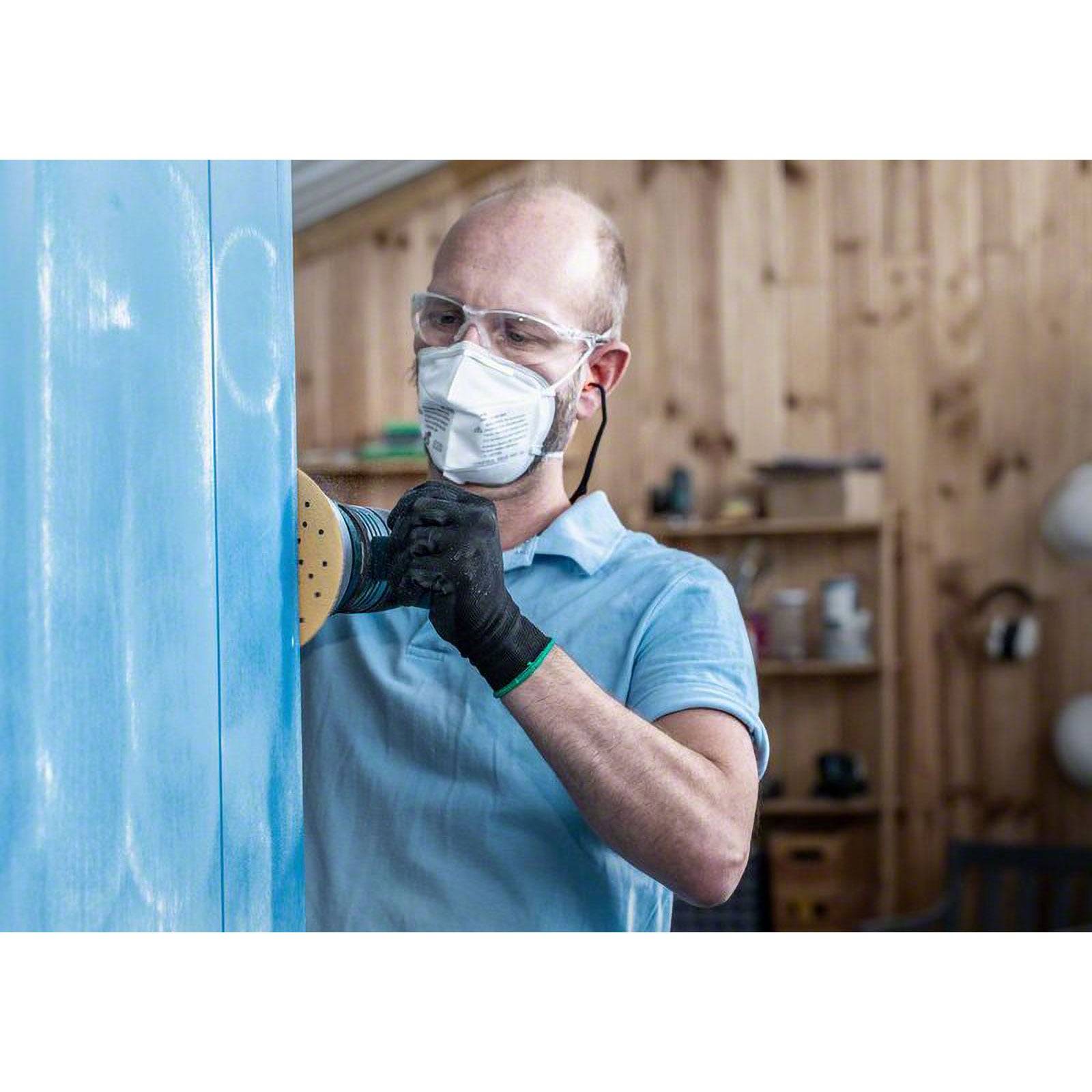 Eine Person trägt Schutzbrille, Maske und Handschuhe und schleift eine blaue Holzoberfläche im Innenbereich und konzentriert sich dabei auf Präzision und Sicherheit.