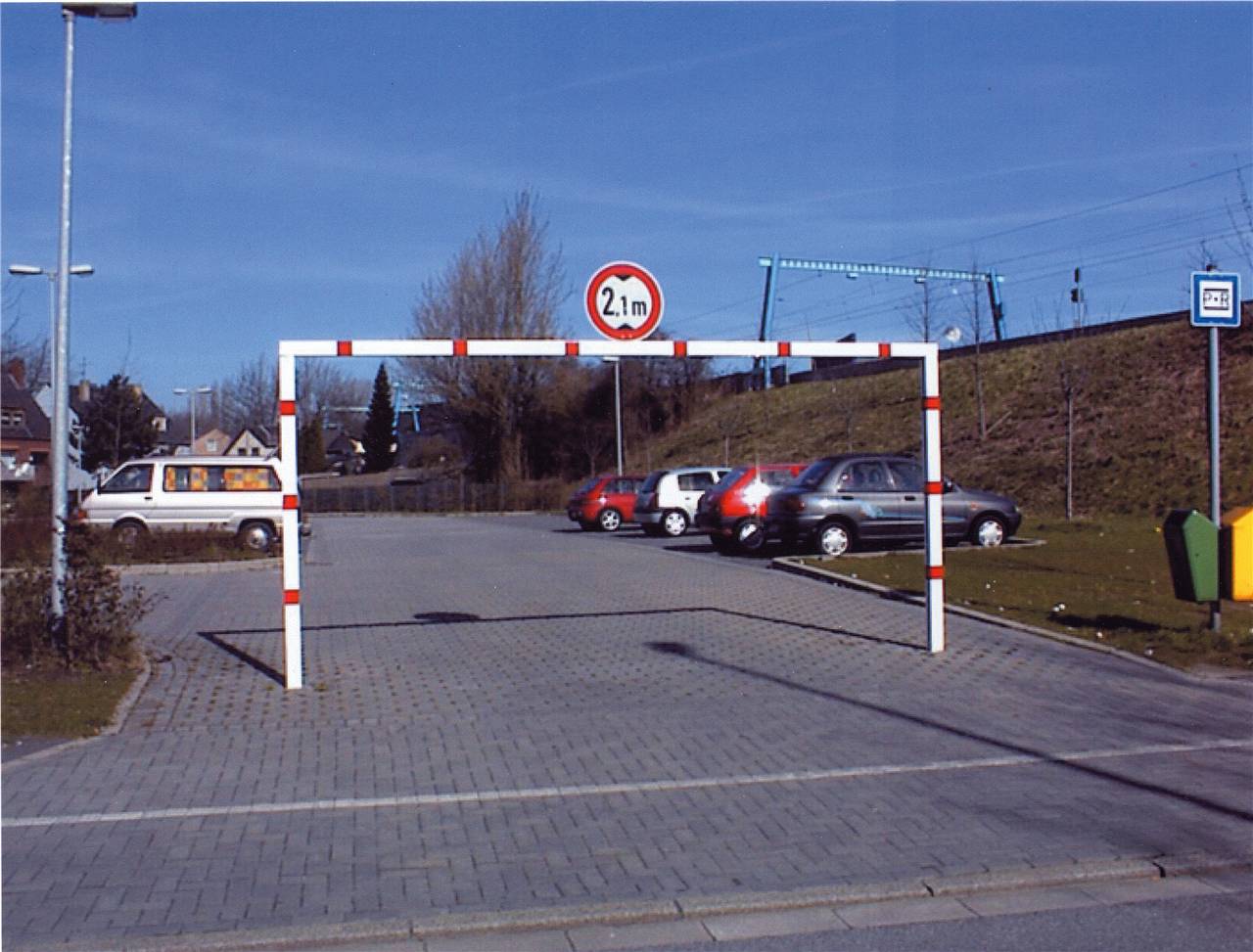 Parkplatz mit einer Durchfahrtssperre, die mit 2,1 m gekennzeichnet ist. Mehrere Autos sind unter dem Höhenbegrenzungsschild und der Durchfahrtssperre geparkt.