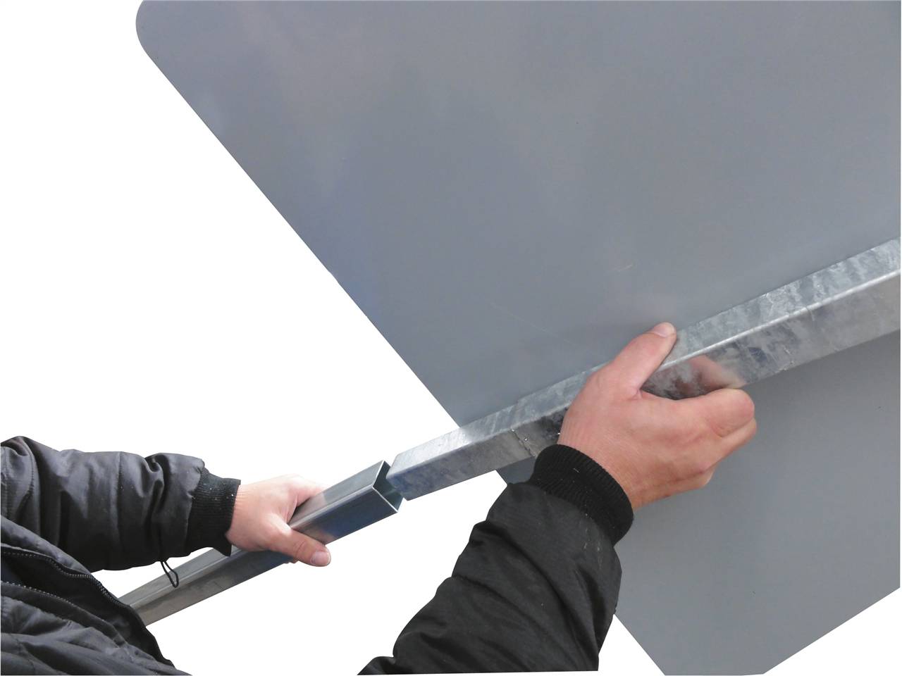 Eine Hand befestigt ein Metallstück an einem grauen Panel, was auf Montage- oder Installationsarbeit hindeutet. Die Person trägt eine schwarze Jacke.