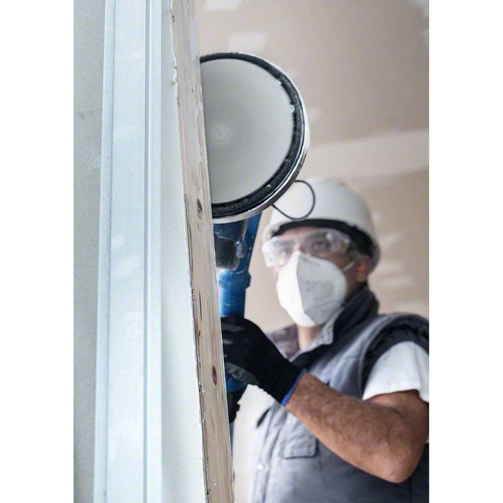 Ein Bauarbeiter schleift Trockenbau mit einer elektrischen Schleifmaschine. Der Arbeiter trägt Schutzausrüstung, einschließlich Schutzhelm, Maske, Schutzbrille und Handschuhen.