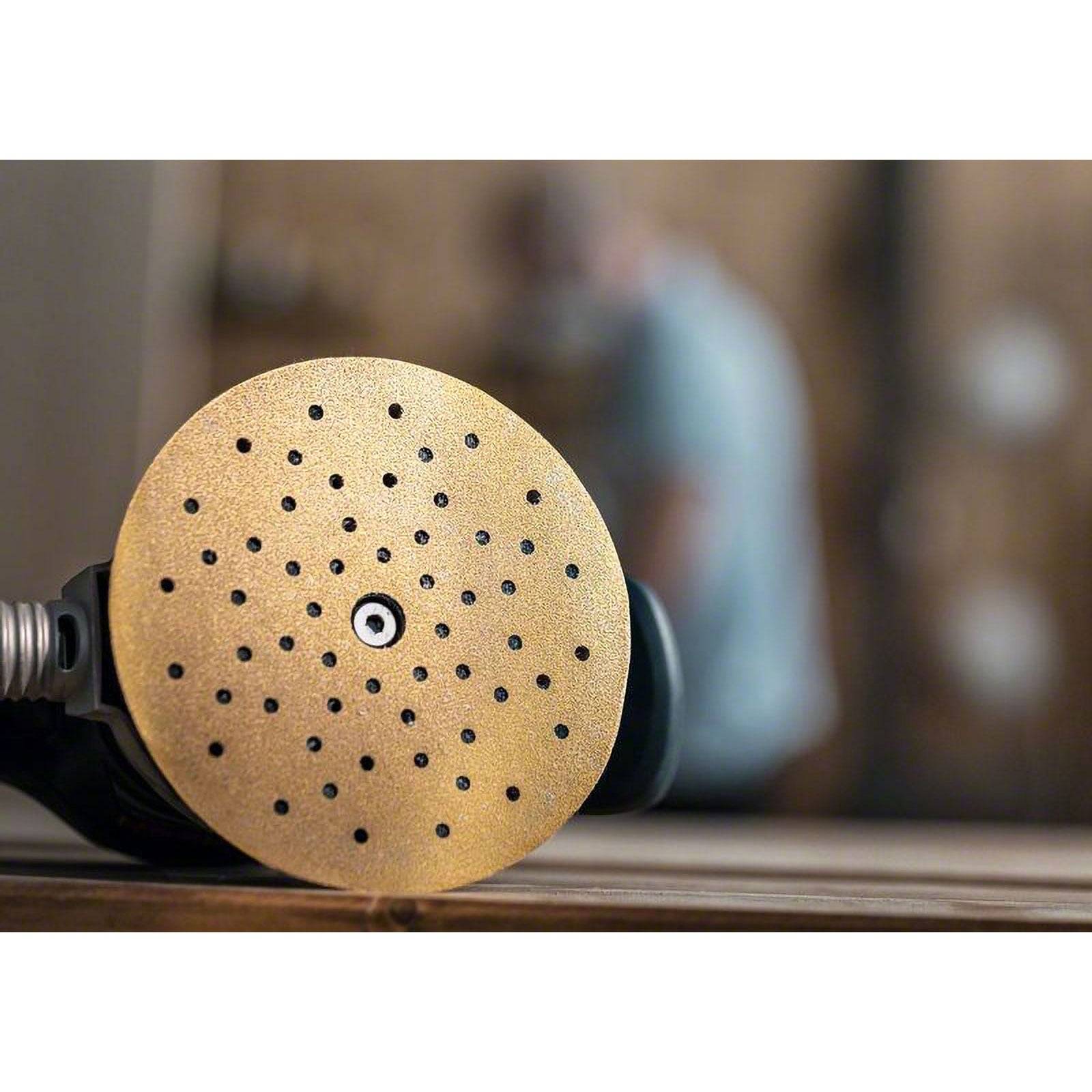 Eine Nahaufnahme einer kreisförmigen Schleifscheibe mit mehreren Löchern liegt auf einer Holzoberfläche, mit einer unscharf im Hintergrund befindlichen Gestalt.