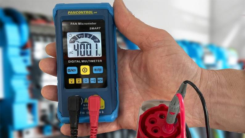 Eine Hand hält ein digitales Multimeter, das 408,1 Volt anzeigt, mit roten und schwarzen Messspitzen, die am unteren Ende angeschlossen sind.