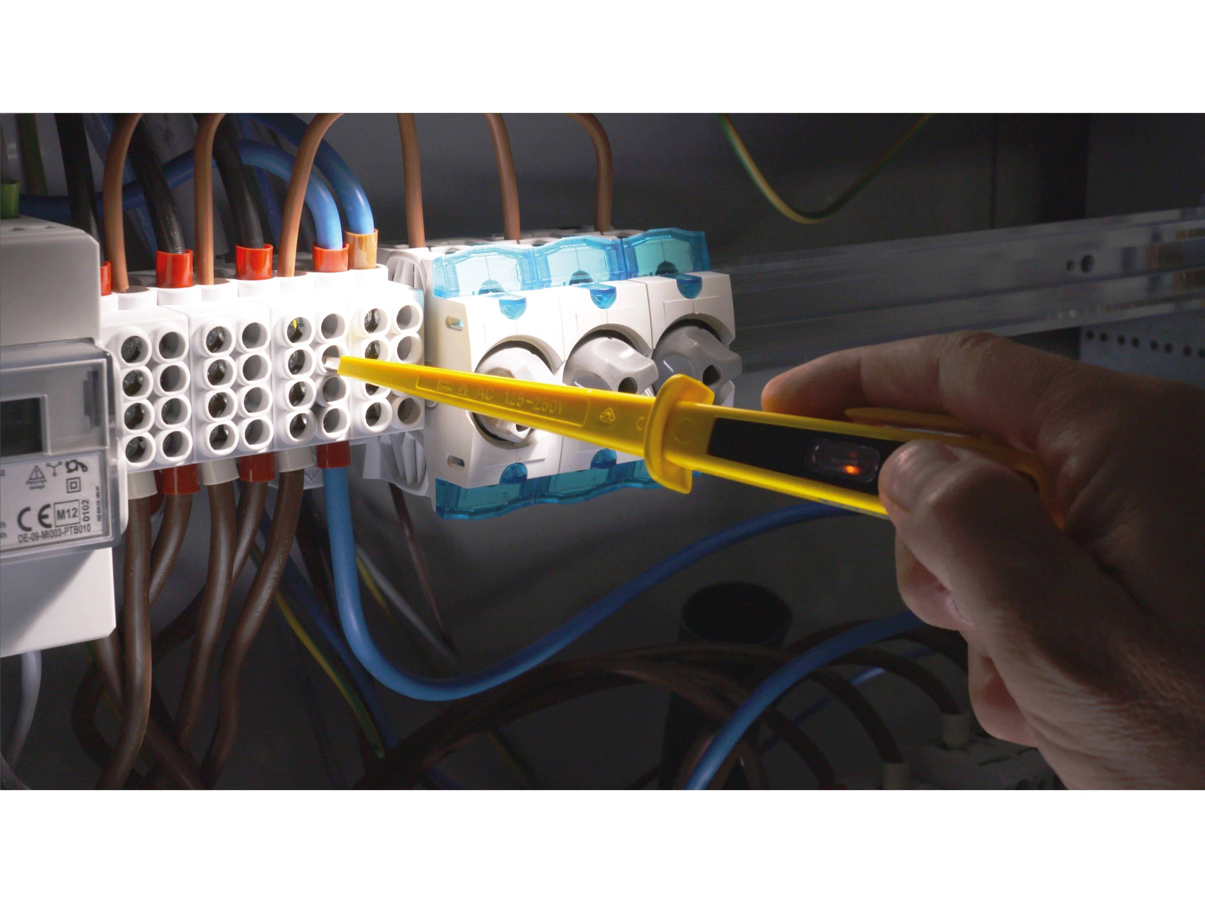 Eine Hand verwendet eine gelbe Spannungsprüfersonde, um einen elektrischen Klemmblock zu überprüfen, umgeben von verschiedenfarbigen Drähten in einer schwach beleuchteten Umgebung.