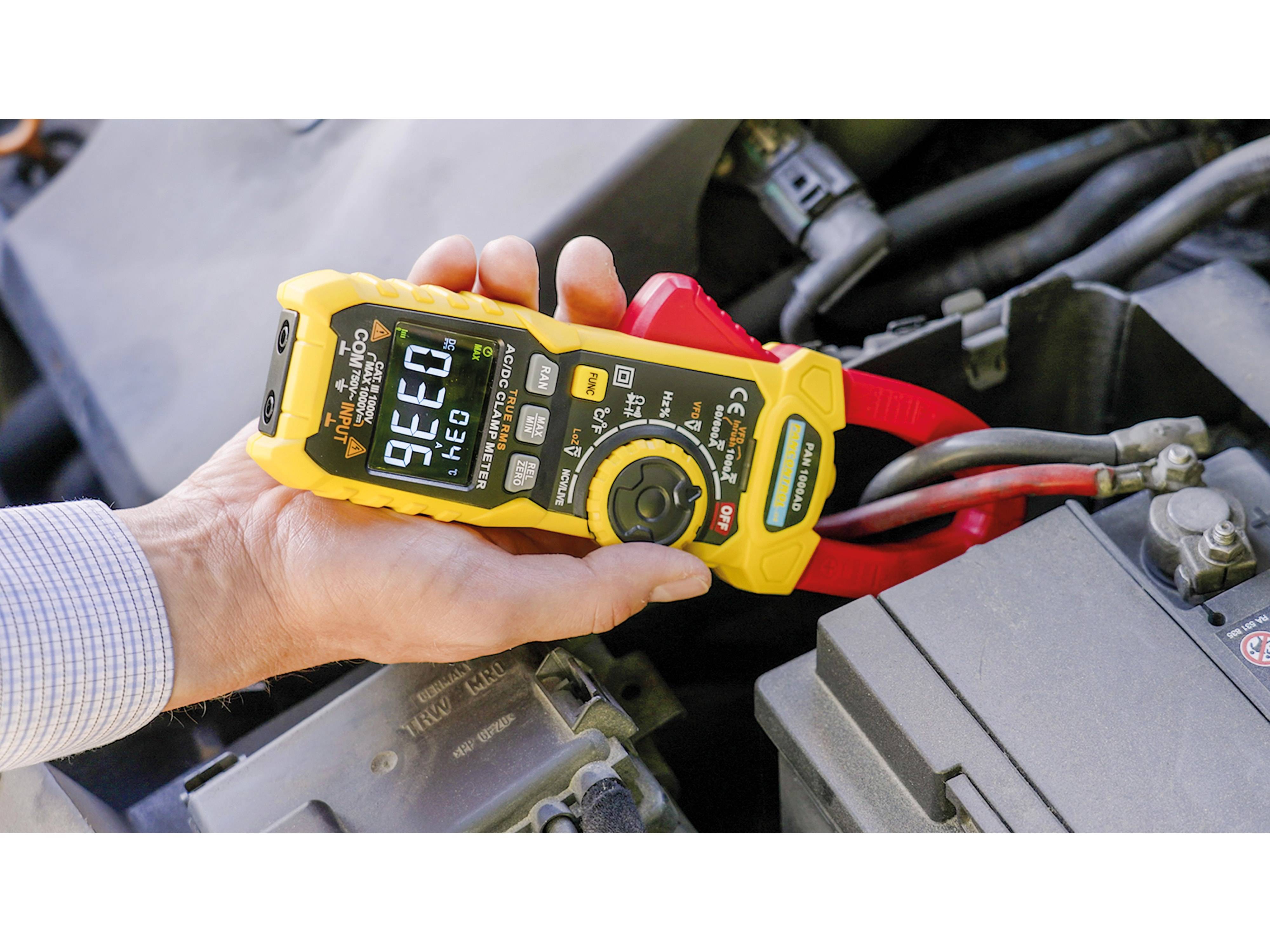 Eine Person verwendet ein gelbes Stromzangenmessgerät, um den elektrischen Strom in einem Automotor zu messen. Das digitale Display zeigt einen Messwert von „9.99